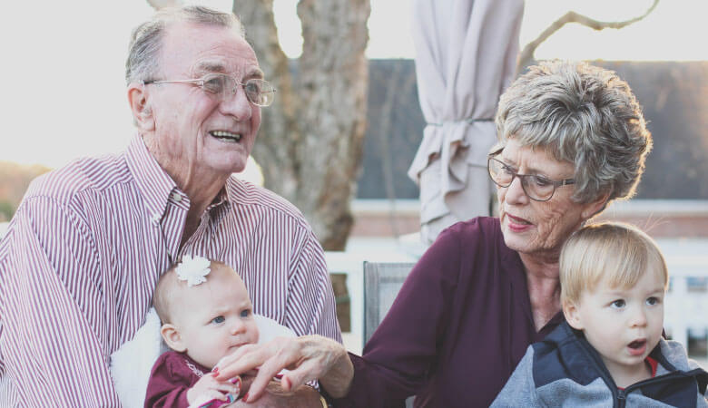 An elderly couple with glasses and purple shirts holds their 2 grandchildren