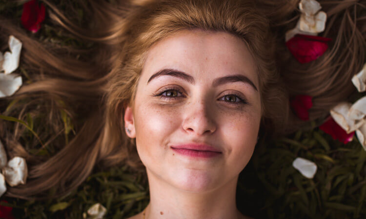 Aerial view of a young woman lying on the grass with rose petals in her hair while smiling with her lips