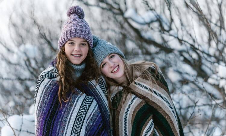 Two teenage girls, one with braces, stand next to each other wrapped in striped blankets and wear beanies in the snow