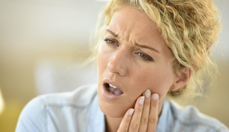 Blonde woman cringes and touches her cheek due to tooth pain from an advanced cavity