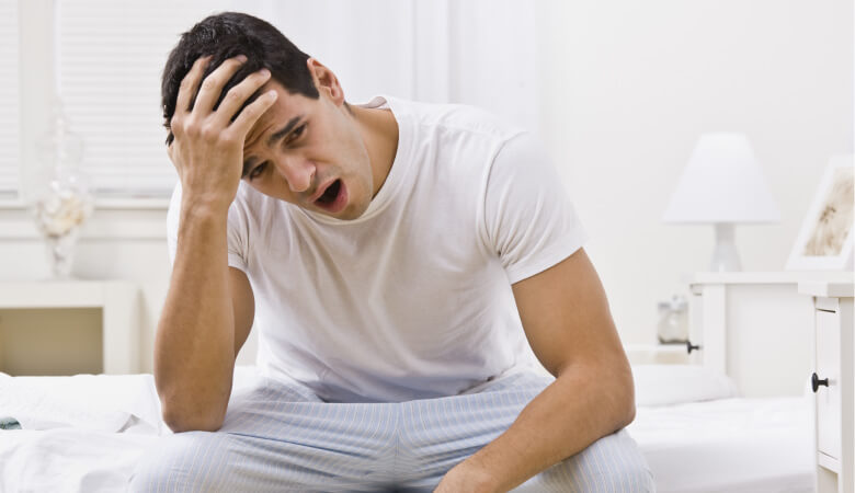 Man sleepily sits on the side of his bed yawning and touching his forehead