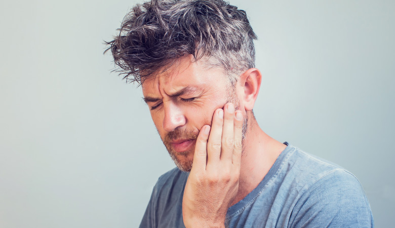 Middle-aged man wearing a gray shirt cringes and touches the side of his cheek due to sinus pain and tooth pain