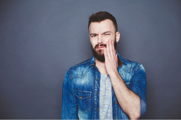 bearded man with a blue jacket holding his jaw with a toothache